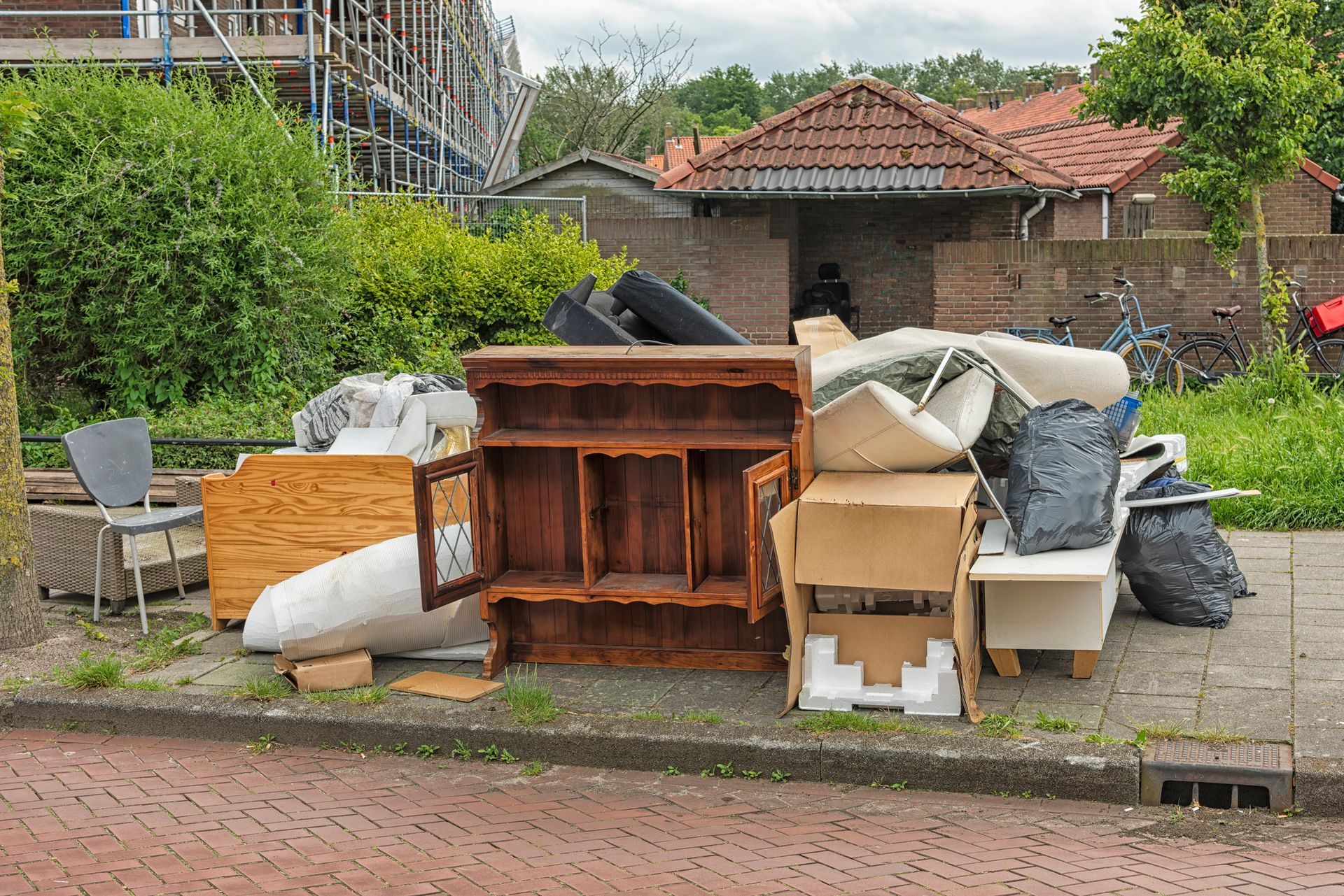 Pile of discarded furniture and trash on a sidewalk next to a brick building and greenery.