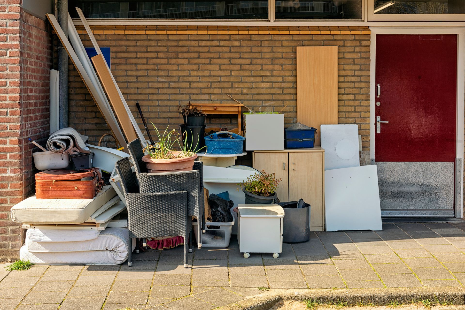 Pile of discarded items: furniture, wood, boxes, and plants next to a brick building with a red door.