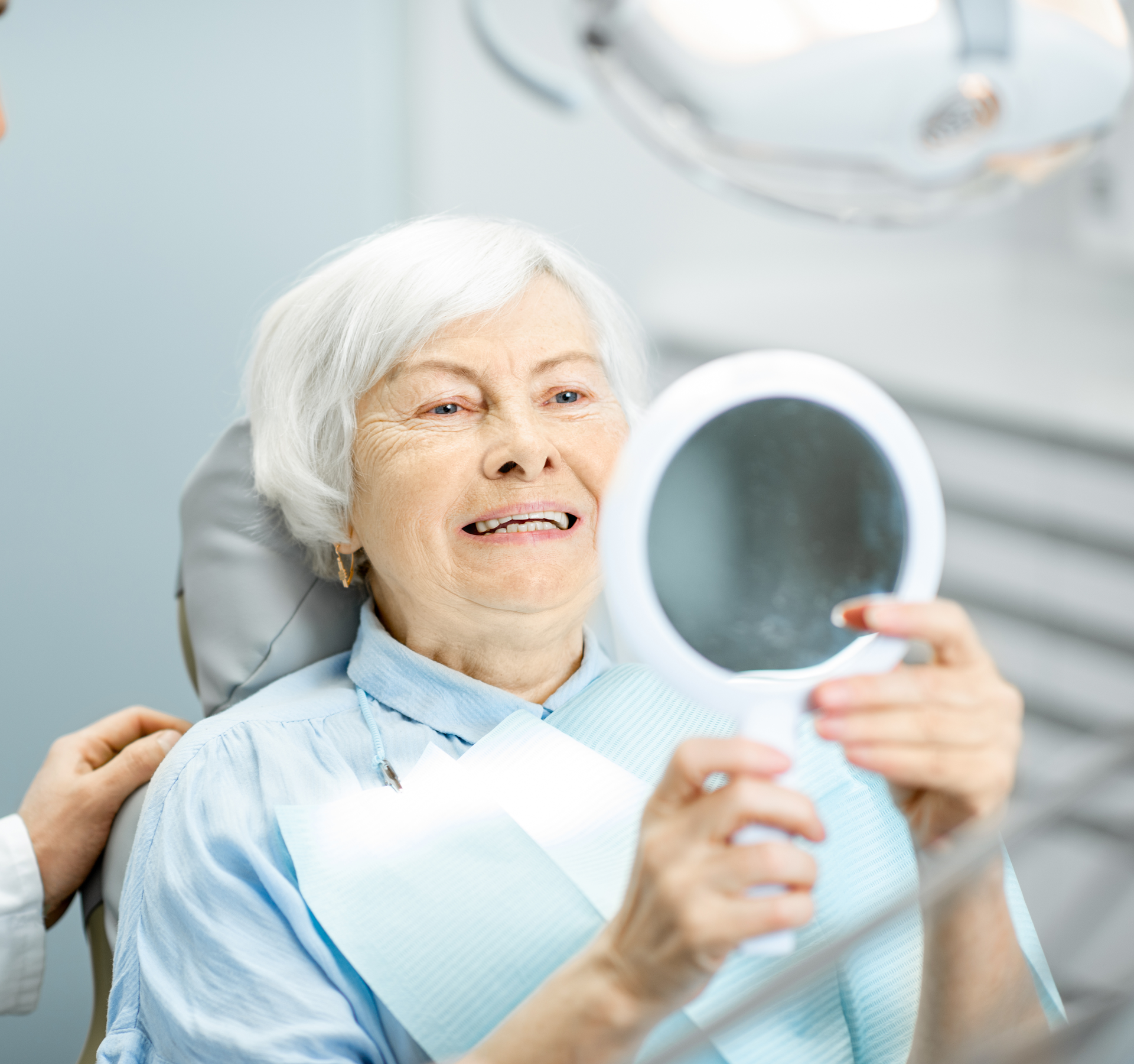 A patient in a blue shirt looks at their teeth in a handheld mirror while seated in a dental chair.