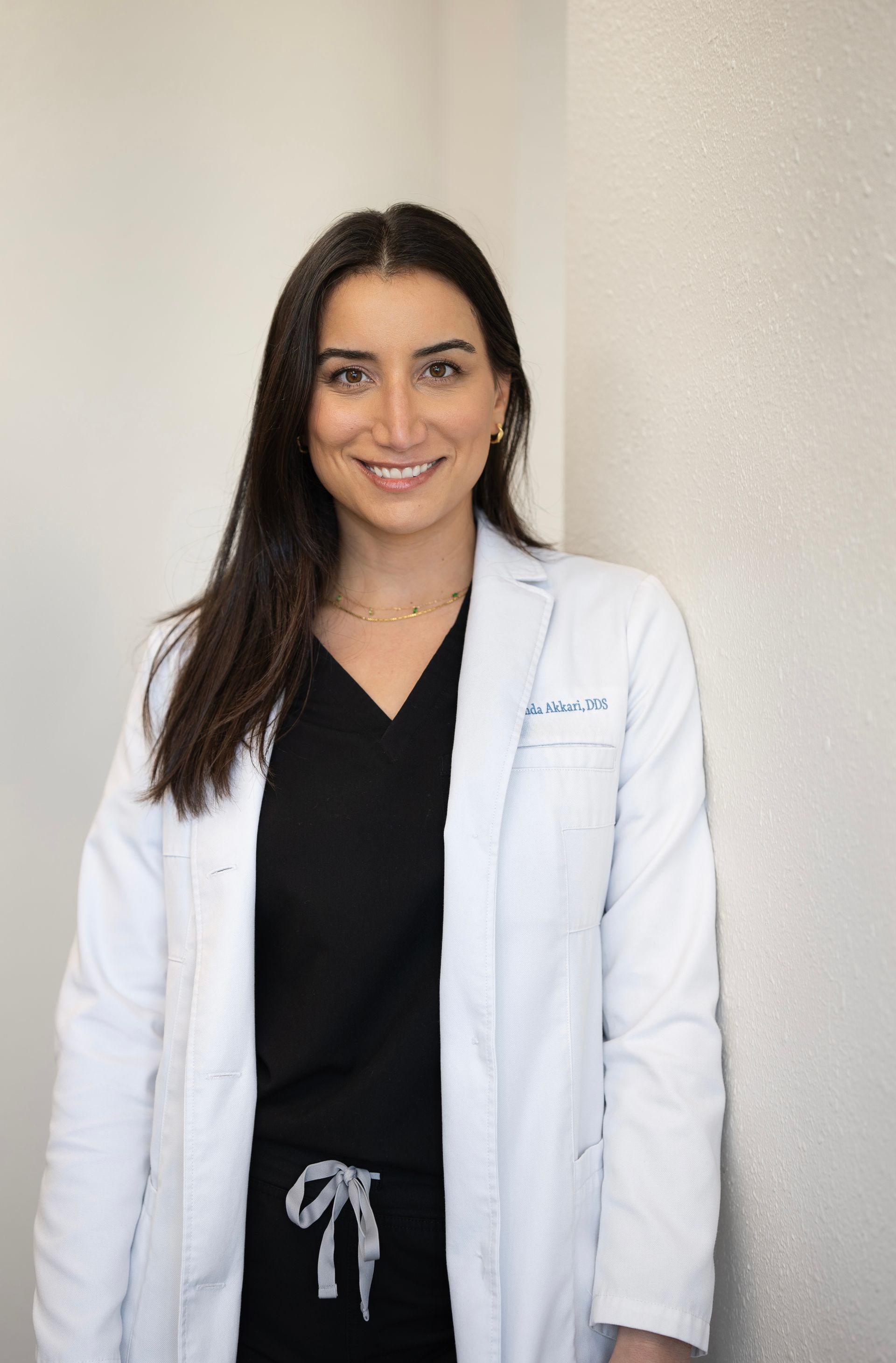 A smiling medical professional with long dark hair, wearing a white lab coat over a black scrub top, leaning against a wall.