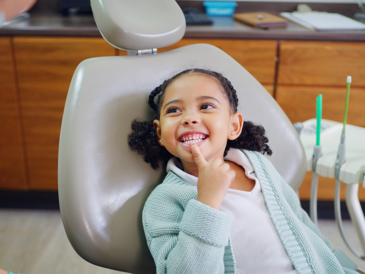 Young child in dentist chair, smiling, pointing to teeth. Dental office setting.