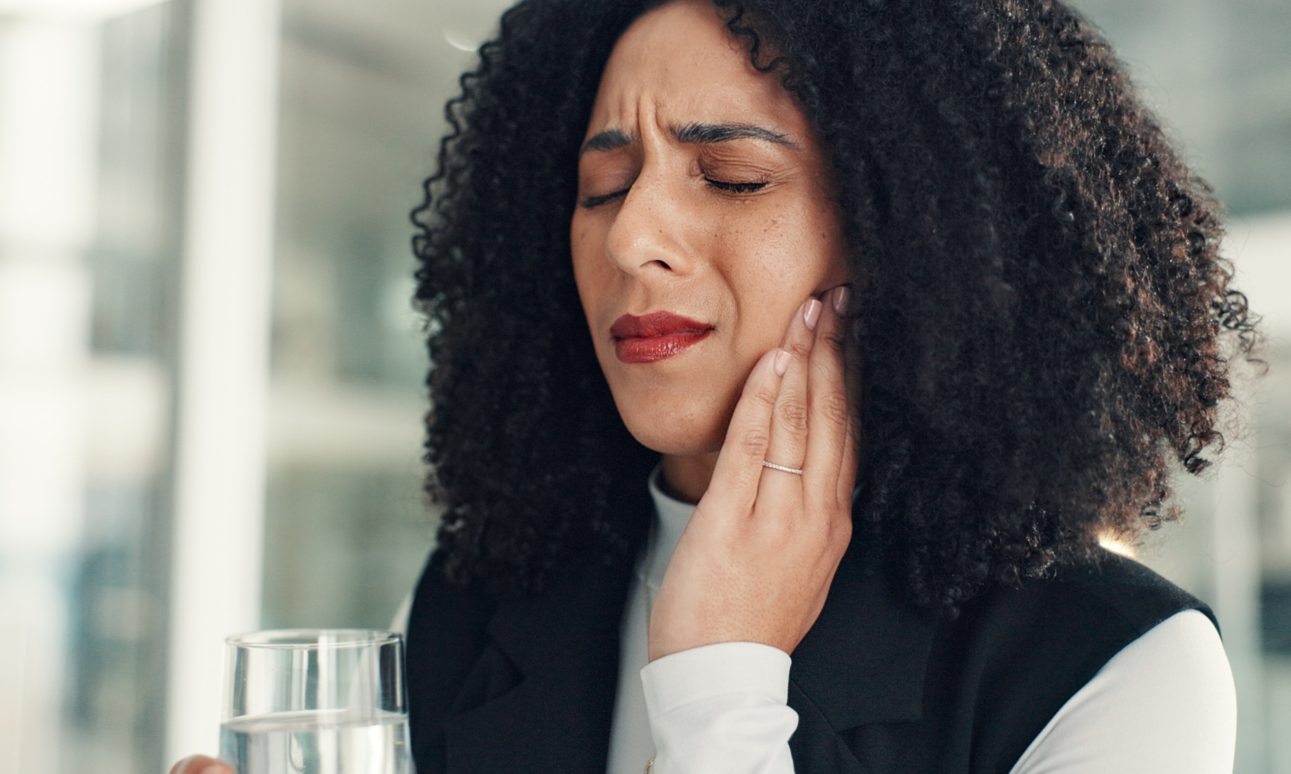 Woman with curly hair holds her jaw, appearing in pain; a glass of water is in the foreground.