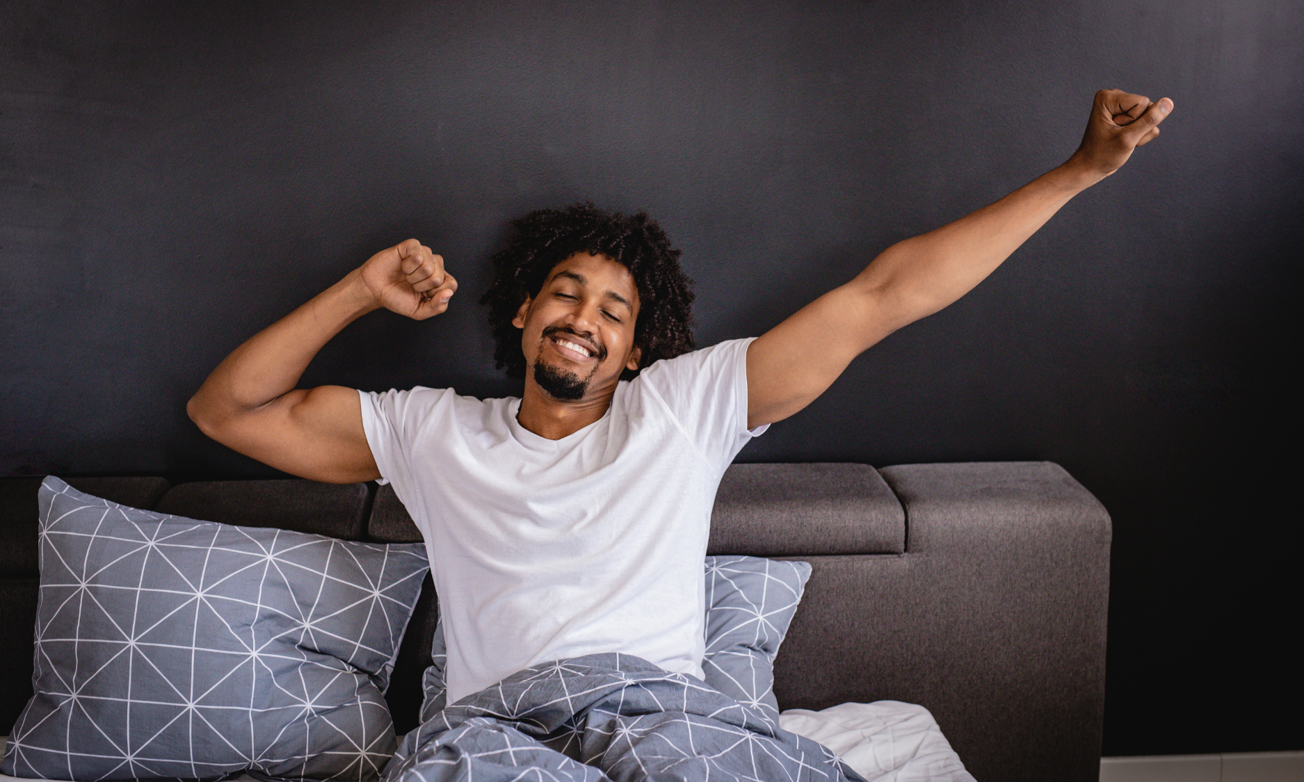 Man sitting up in bed, stretching and smiling. Dark wall, patterned pillow, white shirt, light pants.