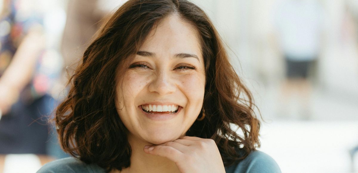 Smiling person with shoulder-length dark hair resting chin on hand outdoors