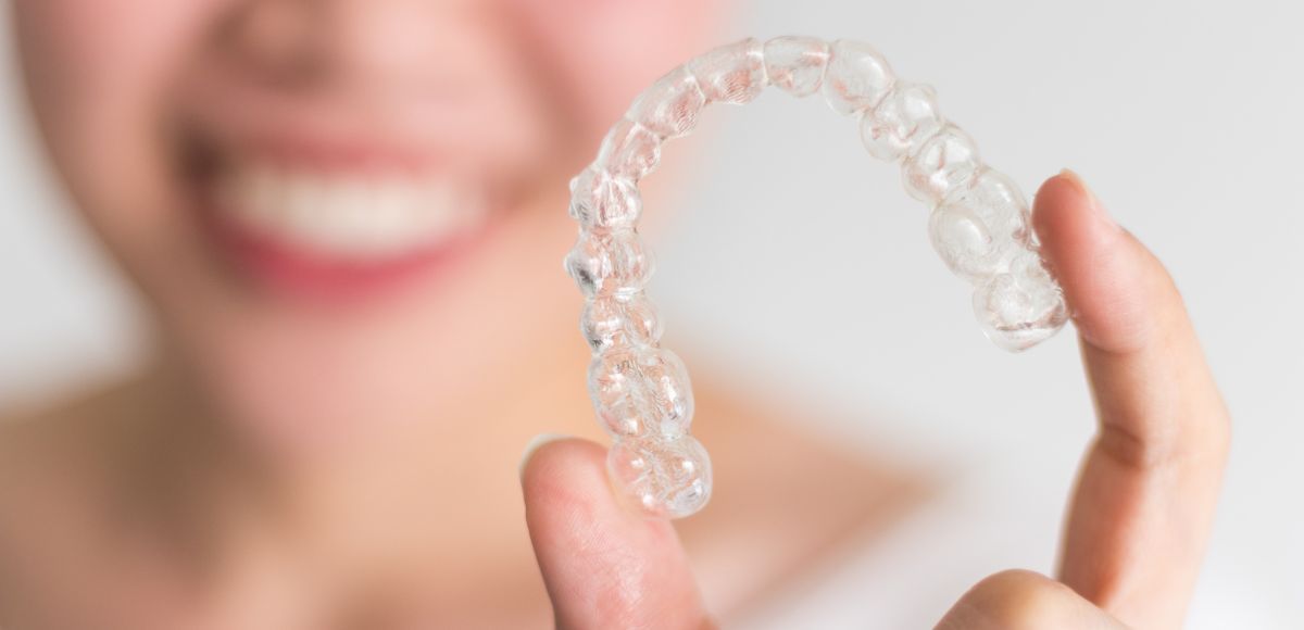 Clear dental aligner held in fingers with a smiling person blurred in the background