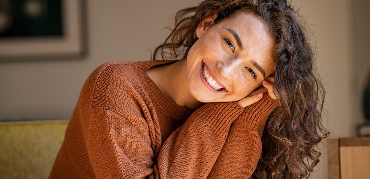 Smiling person in a brown sweater leaning on a wooden table indoors