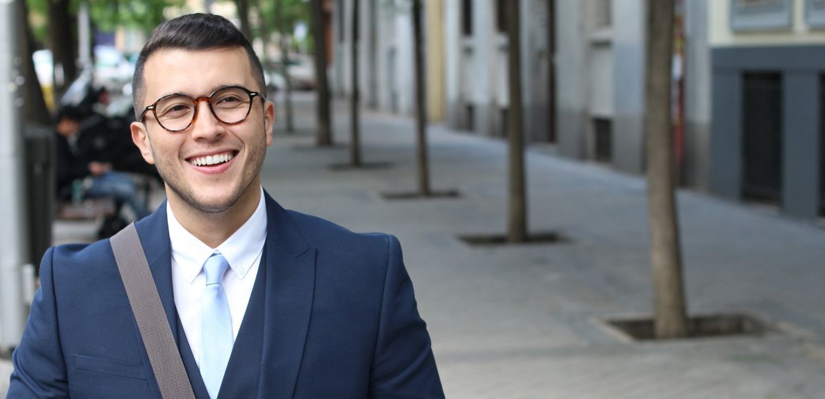 Smiling man in a navy suit and glasses standing outdoors on a city sidewalk