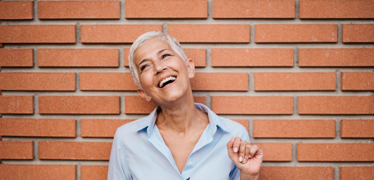 Smiling woman with short white hair posing against a brick wall in a light blue shirt
