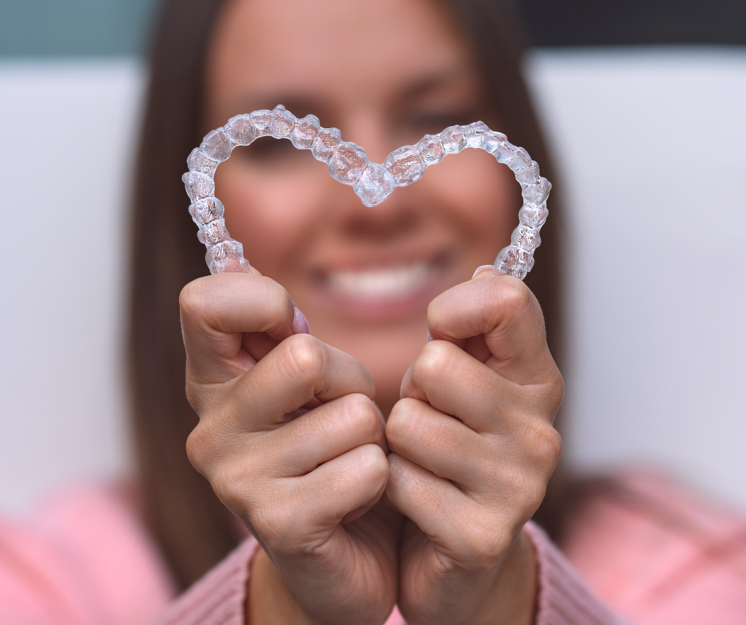 blurred image of a woman in the background with white teeth, holding a heart-shaped invisalign