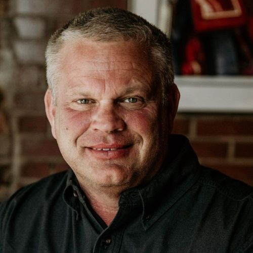 A man in a black shirt is smiling in front of a brick wall.