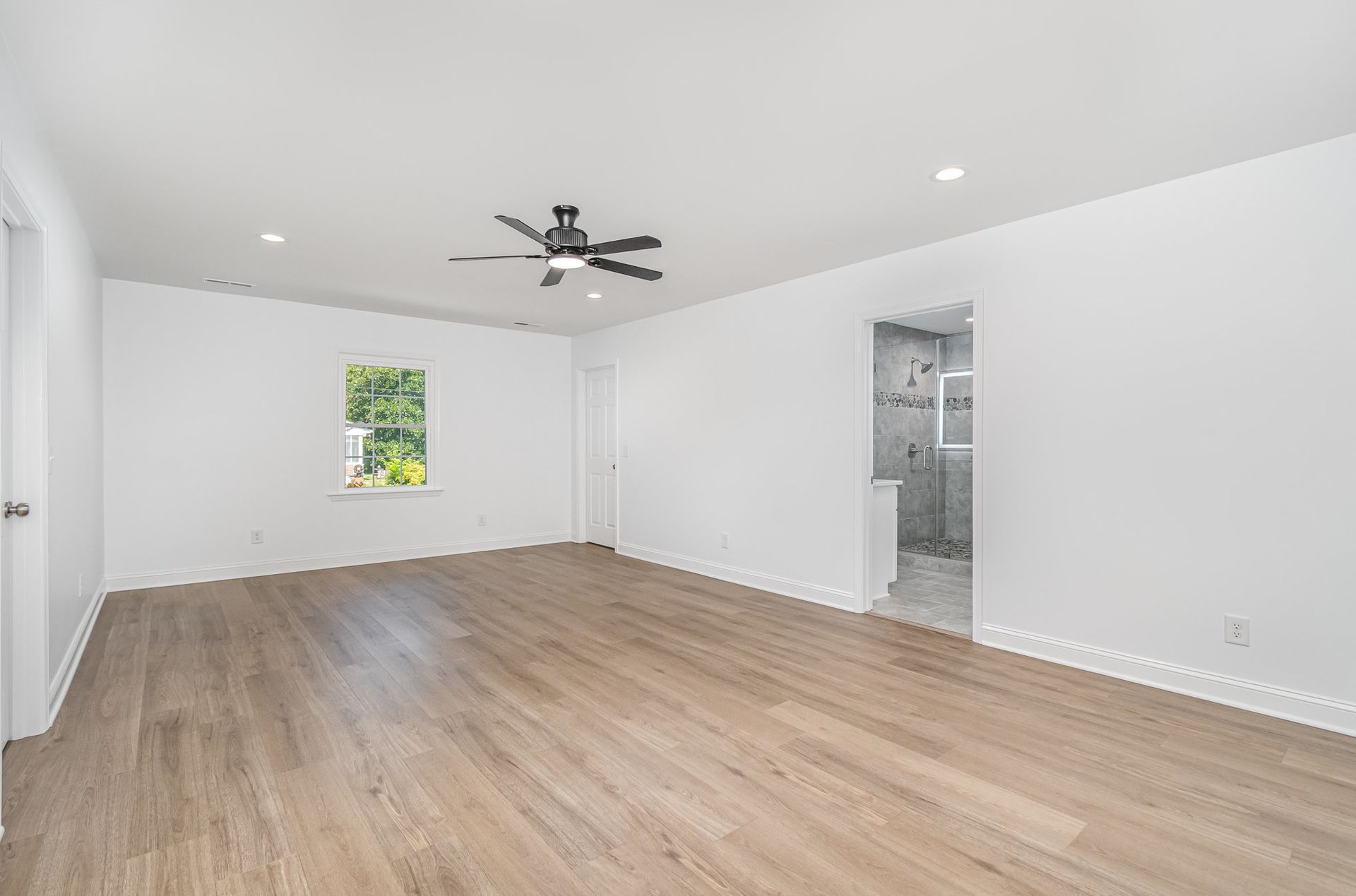 Empty white room with light wood floors, a window, and a bathroom entrance.