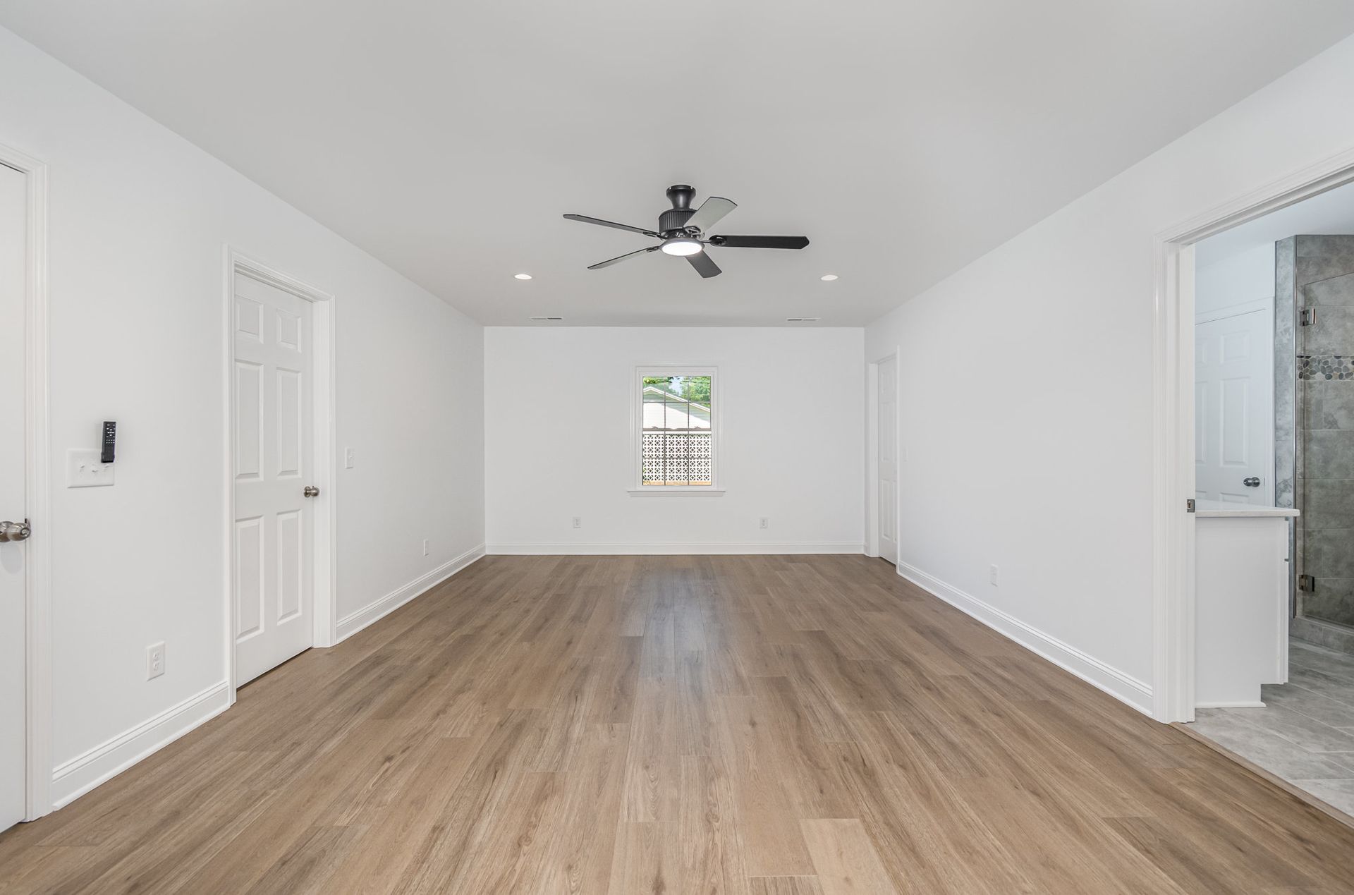 Empty, bright bedroom with white walls, wood-look floors, ceiling fan, and a small window.