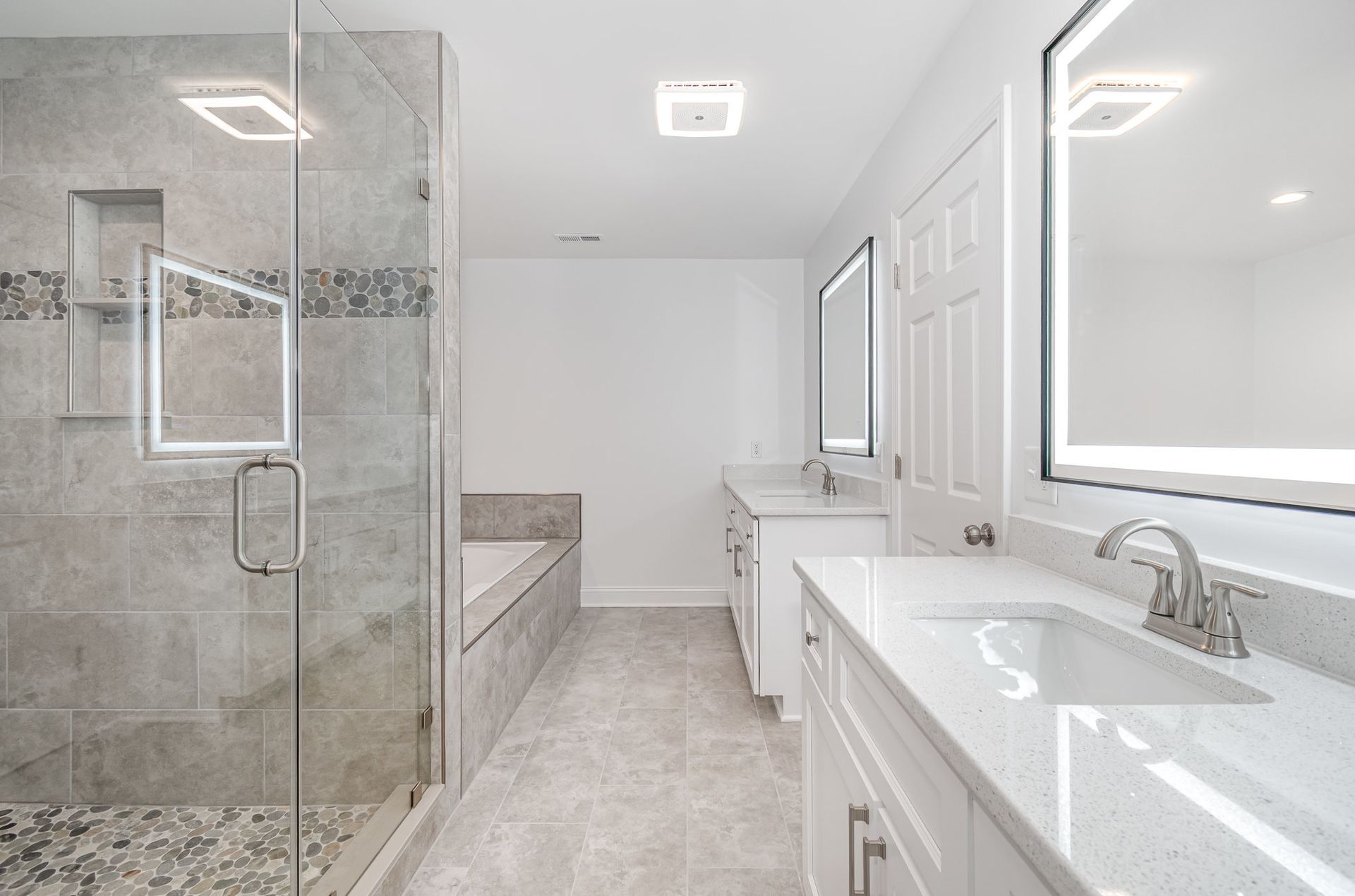 Modern white bathroom with glass shower, double sink, and light-up mirrors. Gray tile floor and walls.