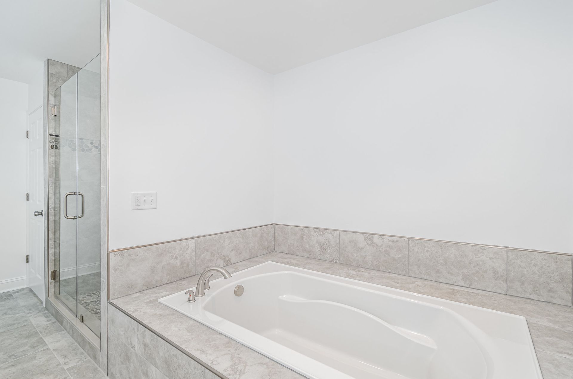 A modern bathroom with a white bathtub and glass shower. Light grey tile and white walls.