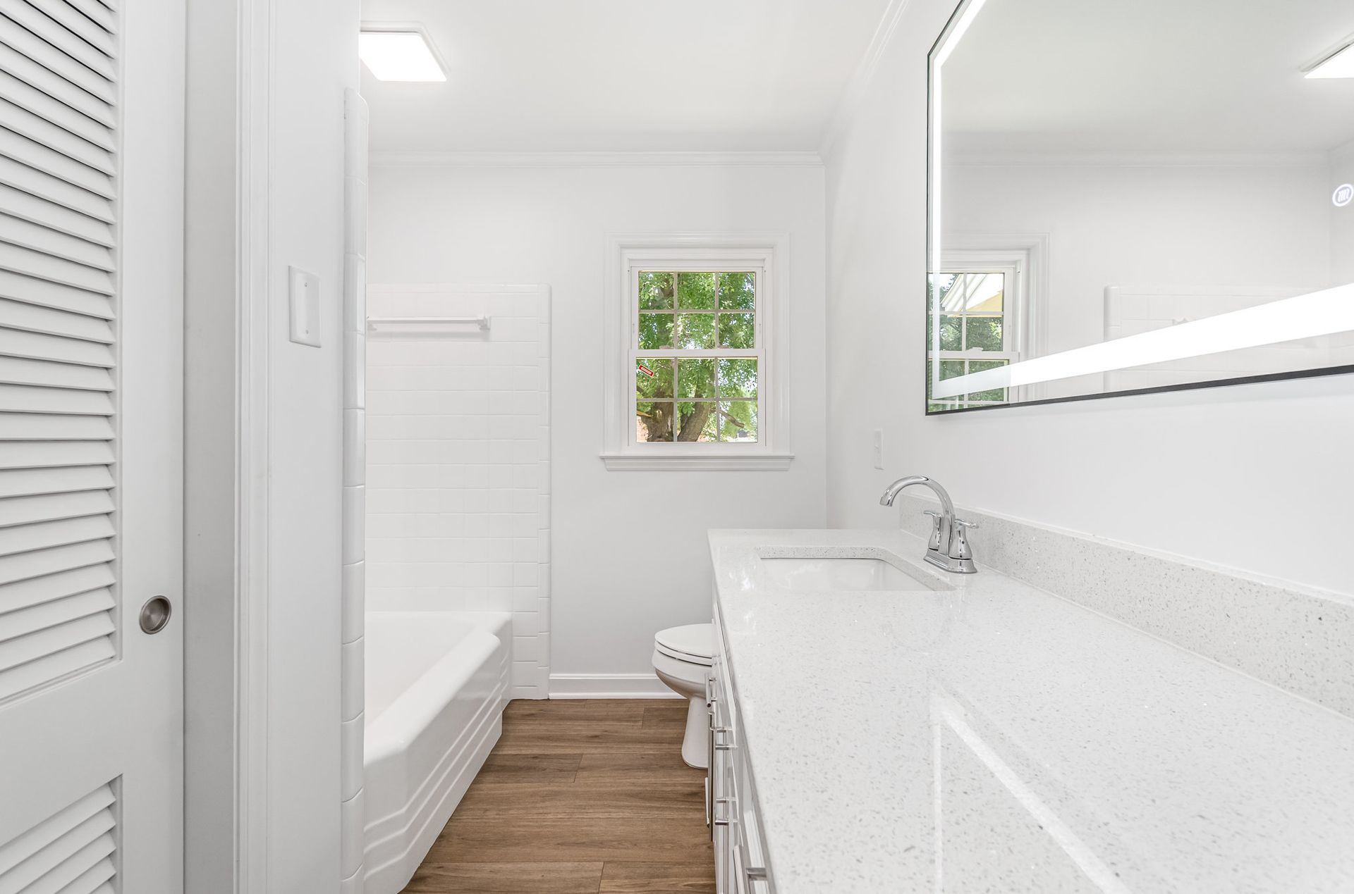 White bathroom with white vanity, tub, and walls. Wood floor. Small window.