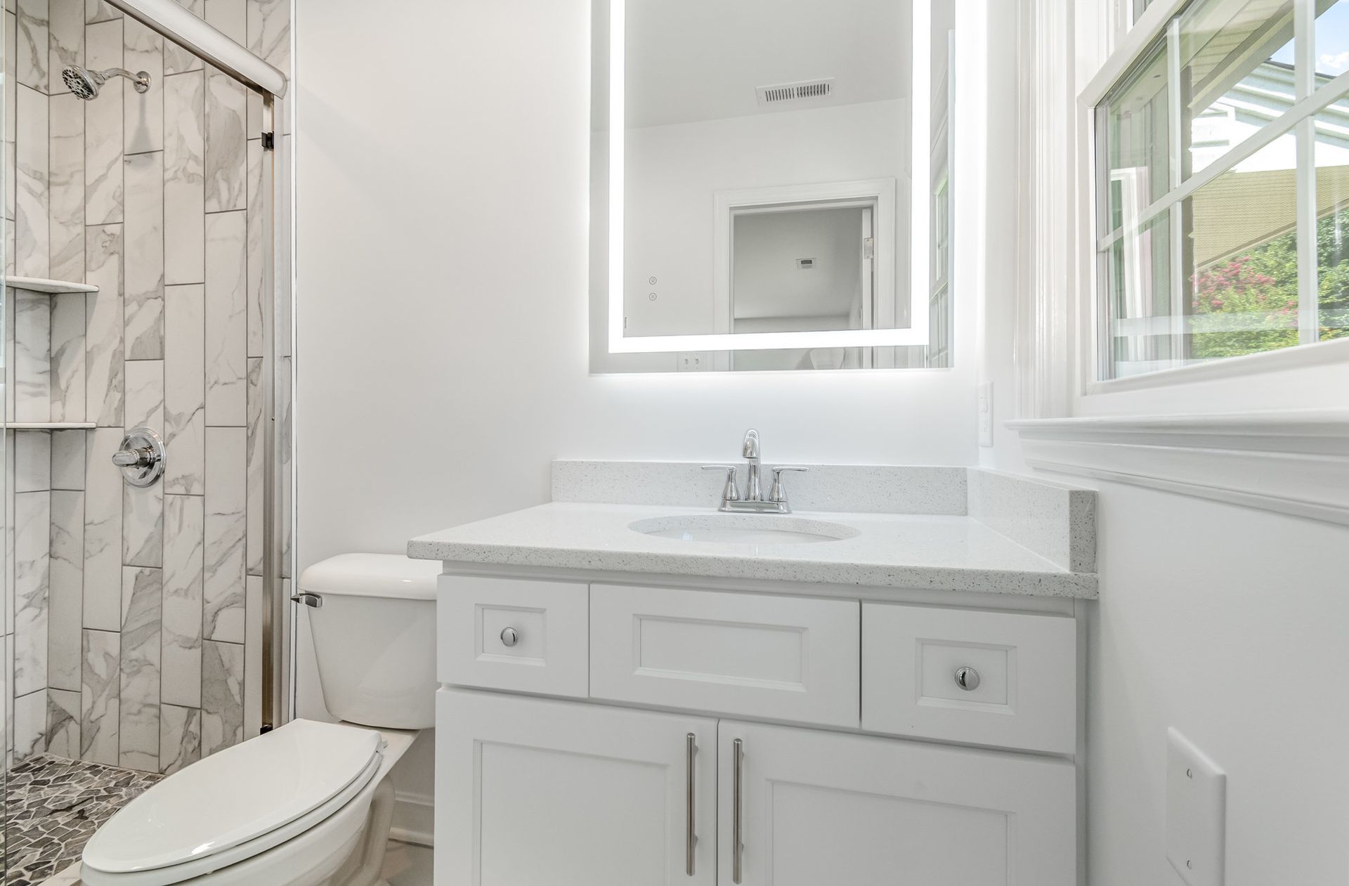 White bathroom with a vanity, toilet, and marble-tiled shower. A large, lit mirror hangs above the vanity.