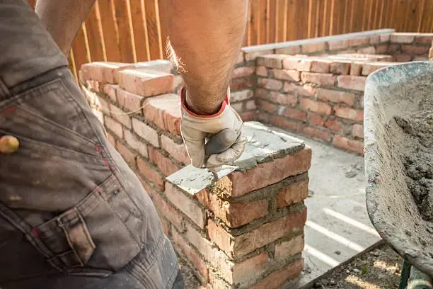 A man is building a brick wall with a wheelbarrow in the background.
