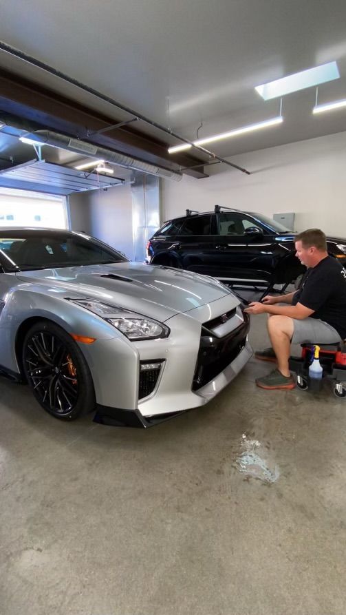 A man is sitting next to a silver sports car in a garage.