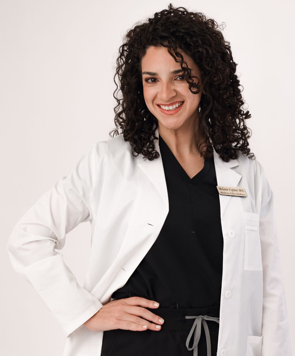 A medical professional in a white coat with a stethoscope around their neck, smiling with arms crossed in an office.