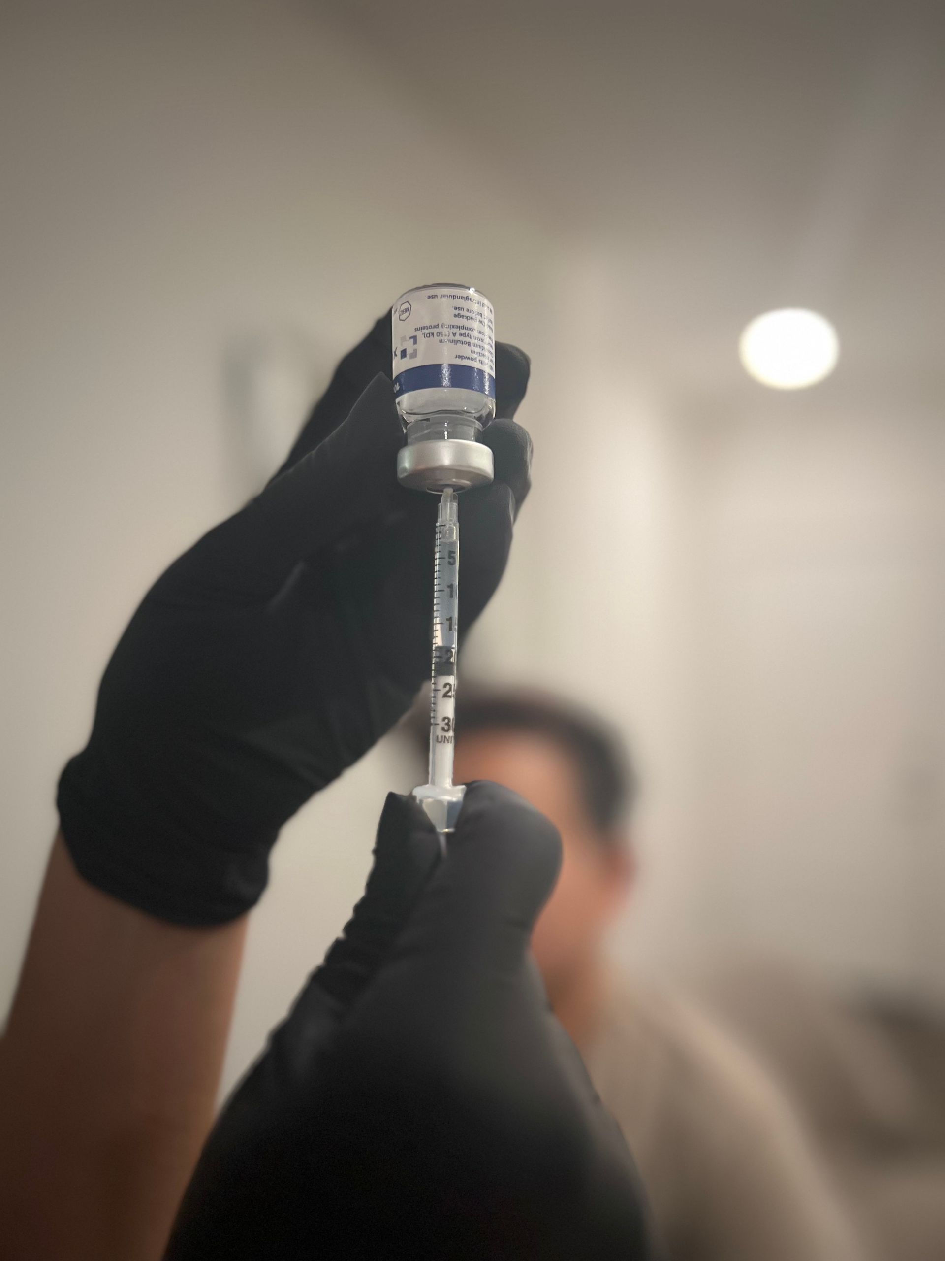A person wearing black medical gloves draws liquid from a small glass vial into a syringe.