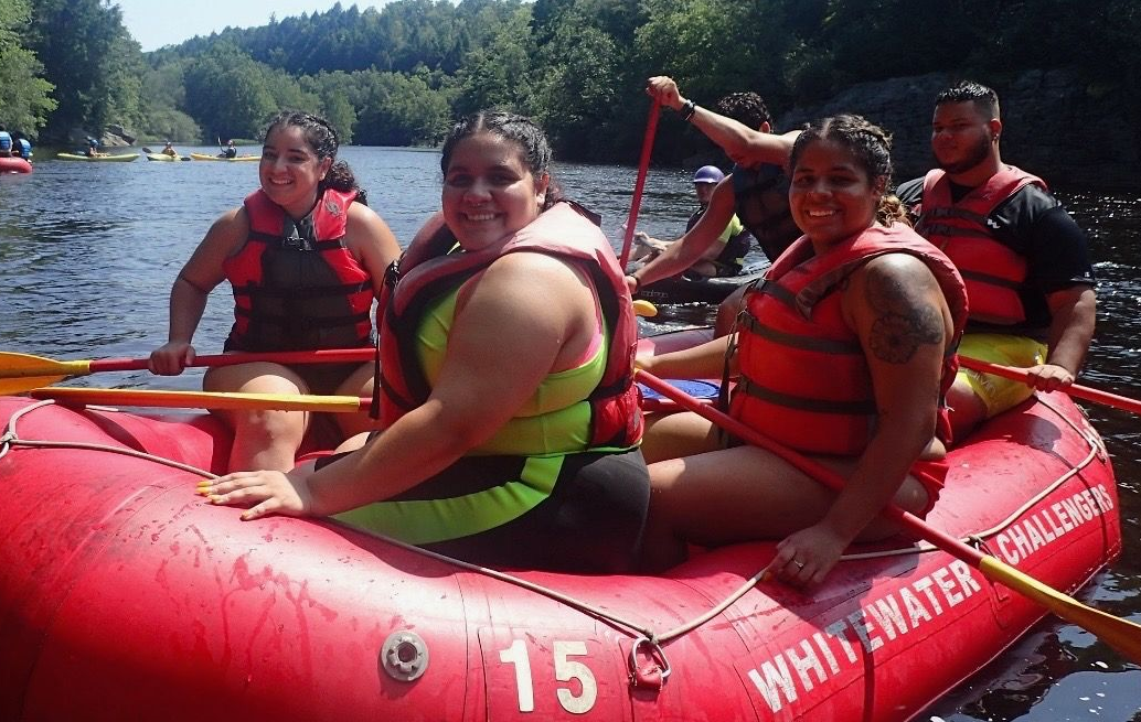 People in life vests and neon gear enjoy whitewater rafting together on a sunny river in a red raft numbered 15.