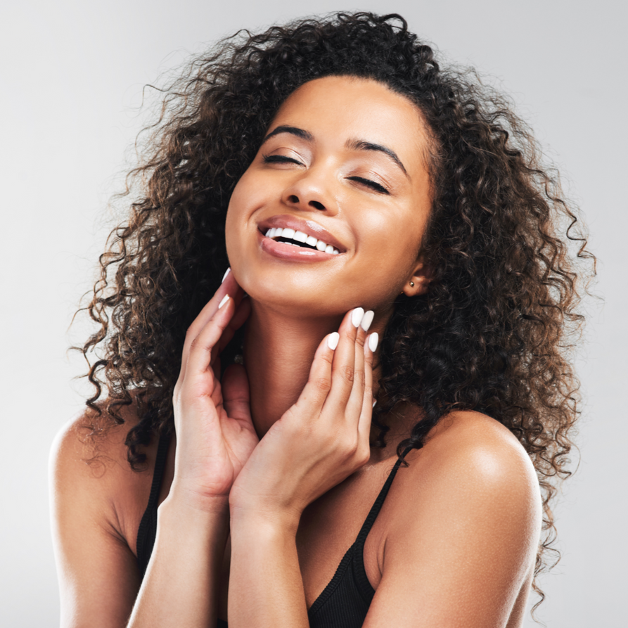 A person with curly hair smiles with their eyes closed, gently touching their neck with both hands against a grey background.