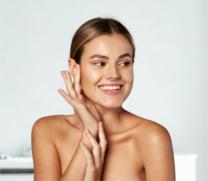 A person with glowing skin smiling while gently touching their cheek with one hand, against a light, neutral background.