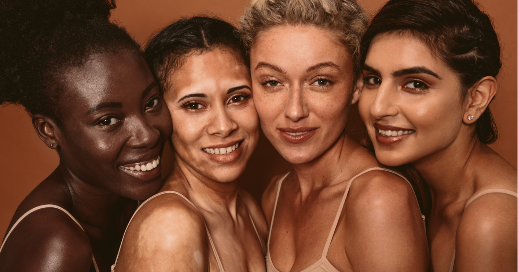 Four people with diverse skin tones and textures are smiling closely together against a solid, warm-toned background.