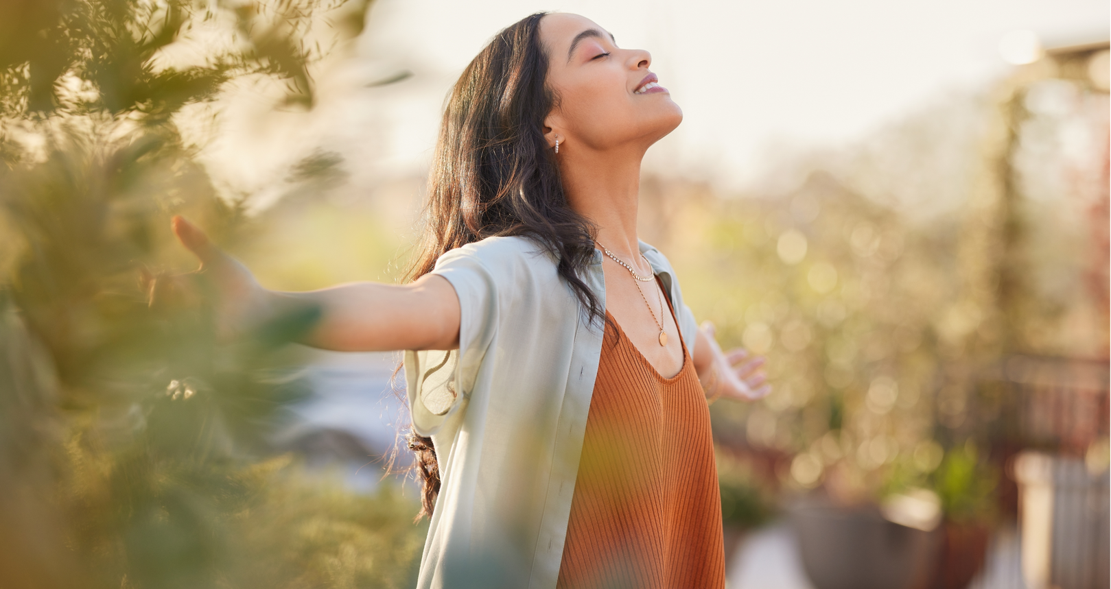 A person with long, dark hair stands outdoors with arms outstretched and eyes closed, appearing peaceful in warm light.