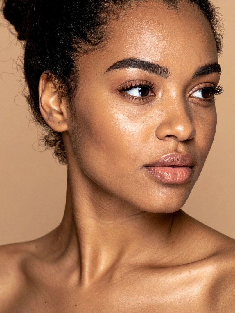A close-up portrait of a person with natural, glowing skin, looking to the side against a neutral beige background.