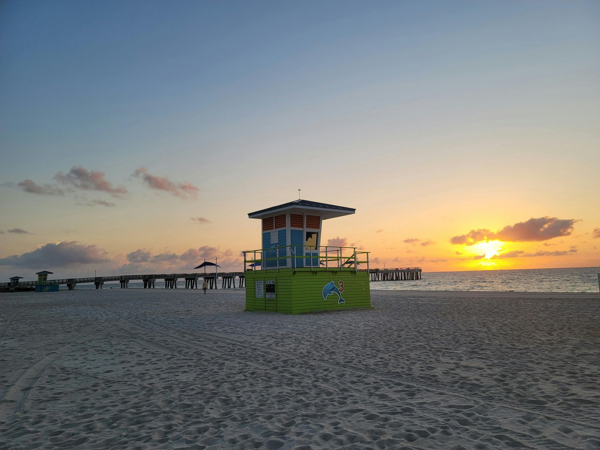 A colorful lifeguard stand on a sandy beach at sunrise, with a pier visible in the distance across the water.