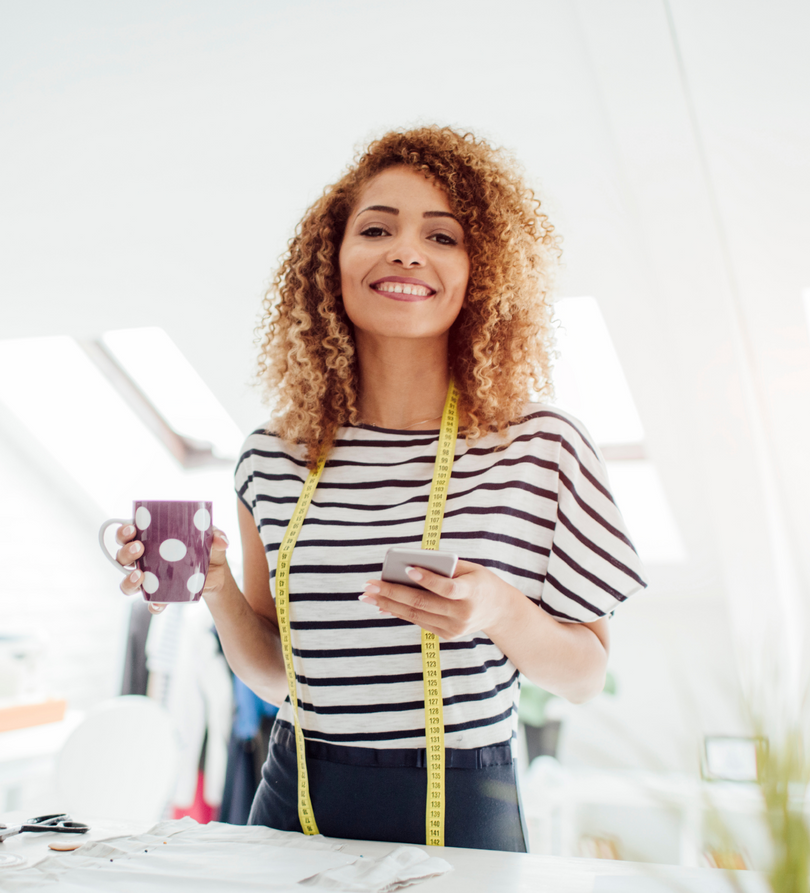A smiling person wearing a striped shirt and measuring tape around their neck holds a mug and a phone in a bright studio.