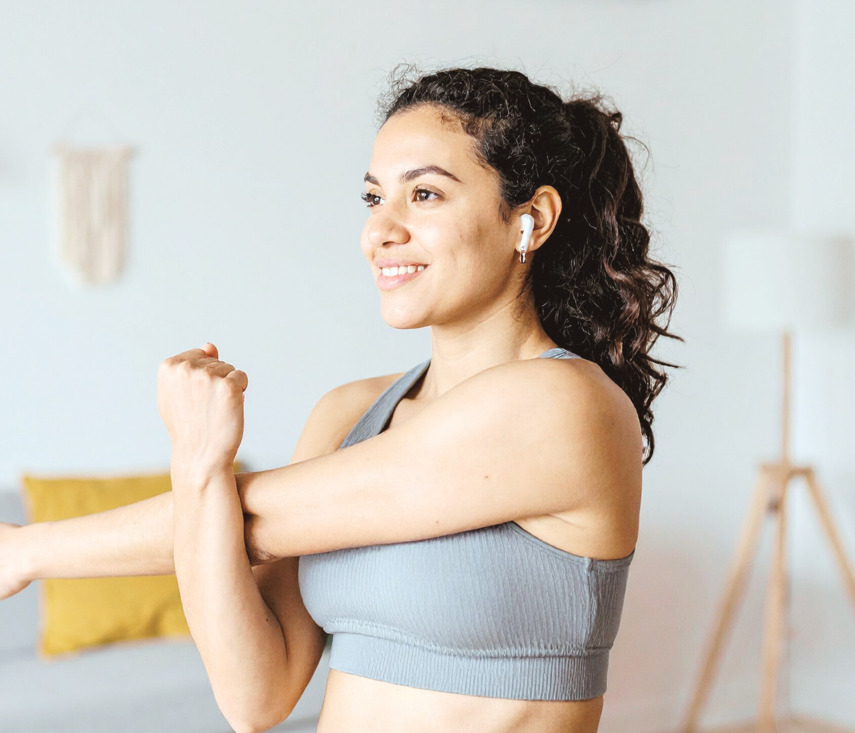 A smiling person in a grey sports bra stands in a bright room, performing a cross-body arm stretch.