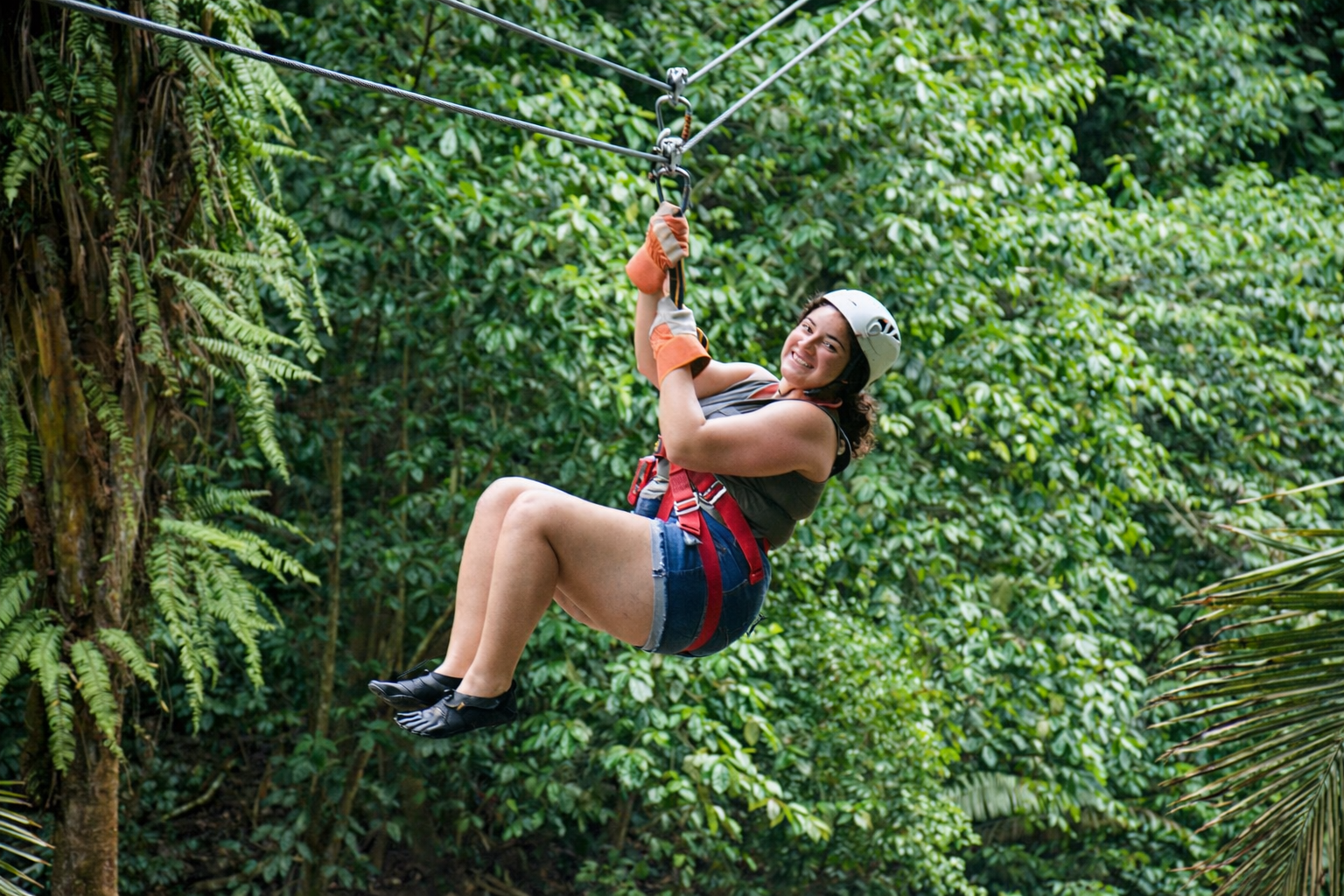 A person wearing a white helmet and harness zips along a cable through a lush, green forest.