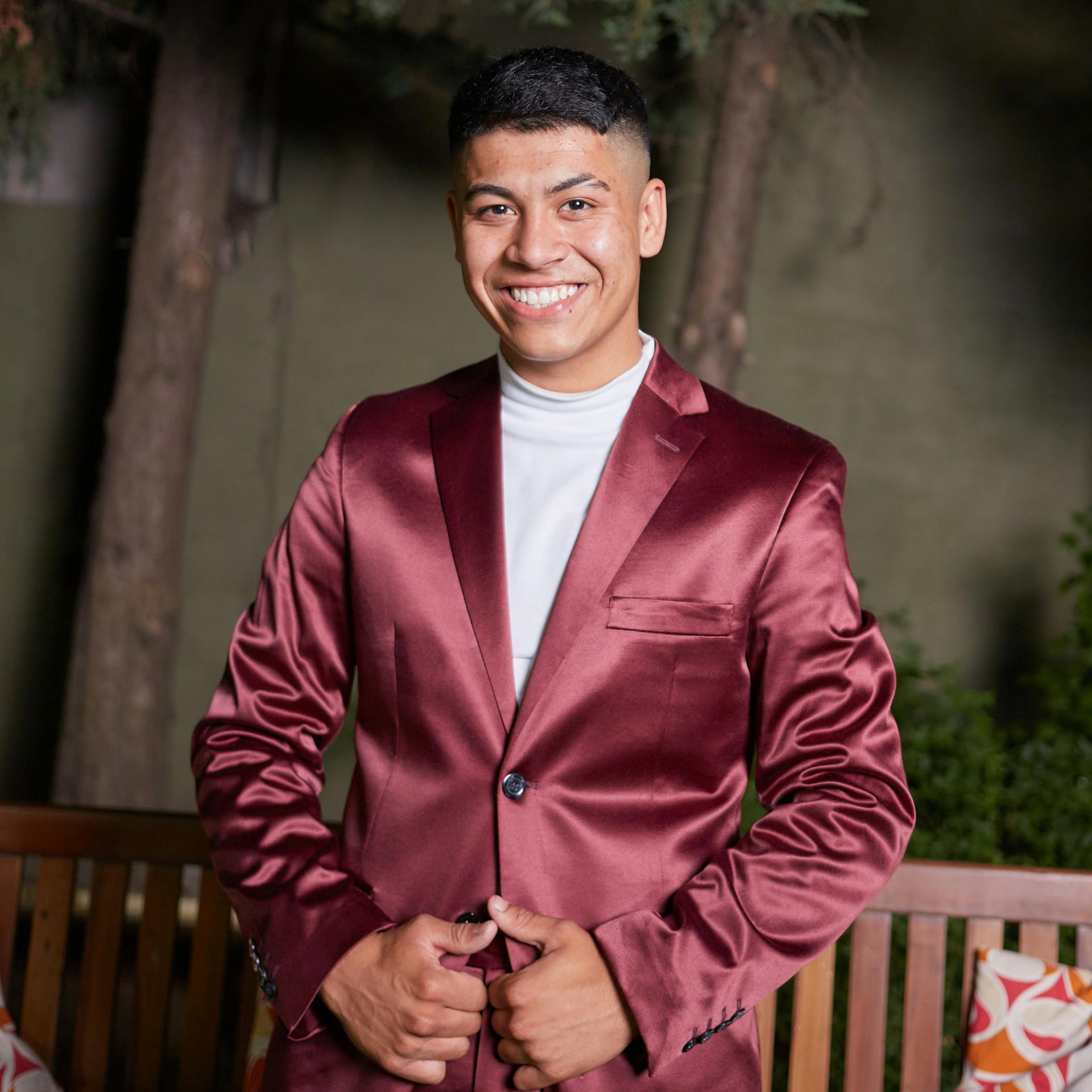 A smiling person wears a maroon satin blazer and white turtleneck, posing in front of a wooden bench and trees.