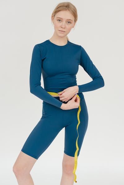 A person in a blue long-sleeved top and biker shorts holds a yellow measuring tape around their waist against a white wall.