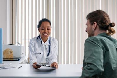 A smiling medical professional in a white coat holds a clipboard while speaking with a patient in an office.