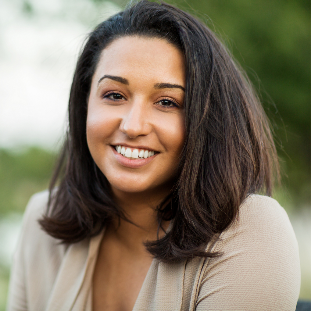A smiling person with shoulder-length dark hair wearing a beige top, set against a softly blurred outdoor green background.