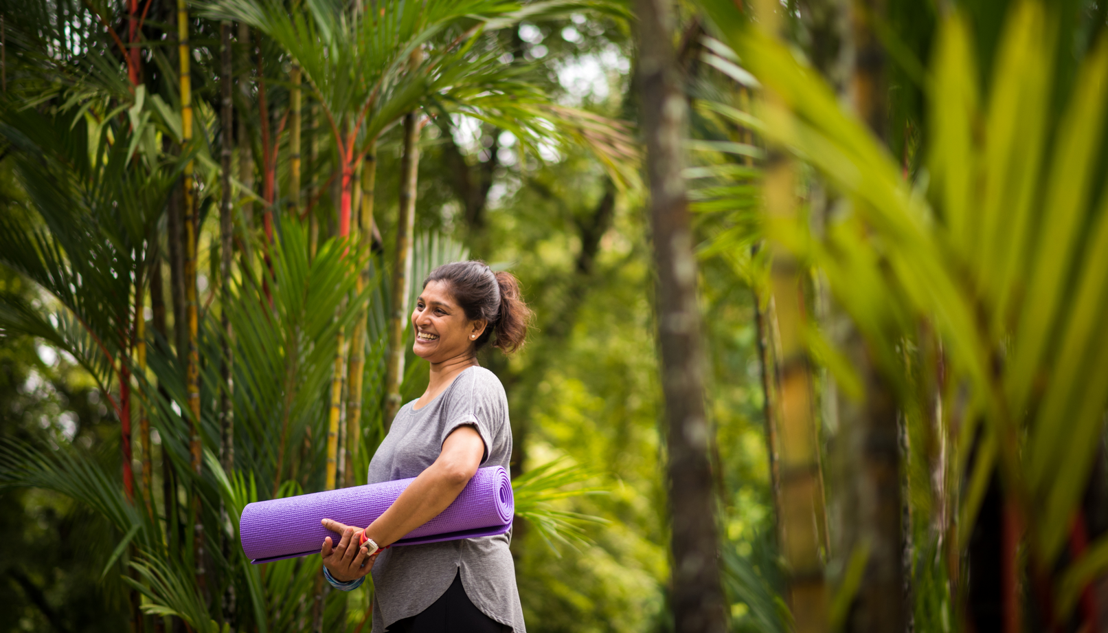 A smiling person stands in a lush, green forest holding a rolled-up purple yoga mat under their arm.
