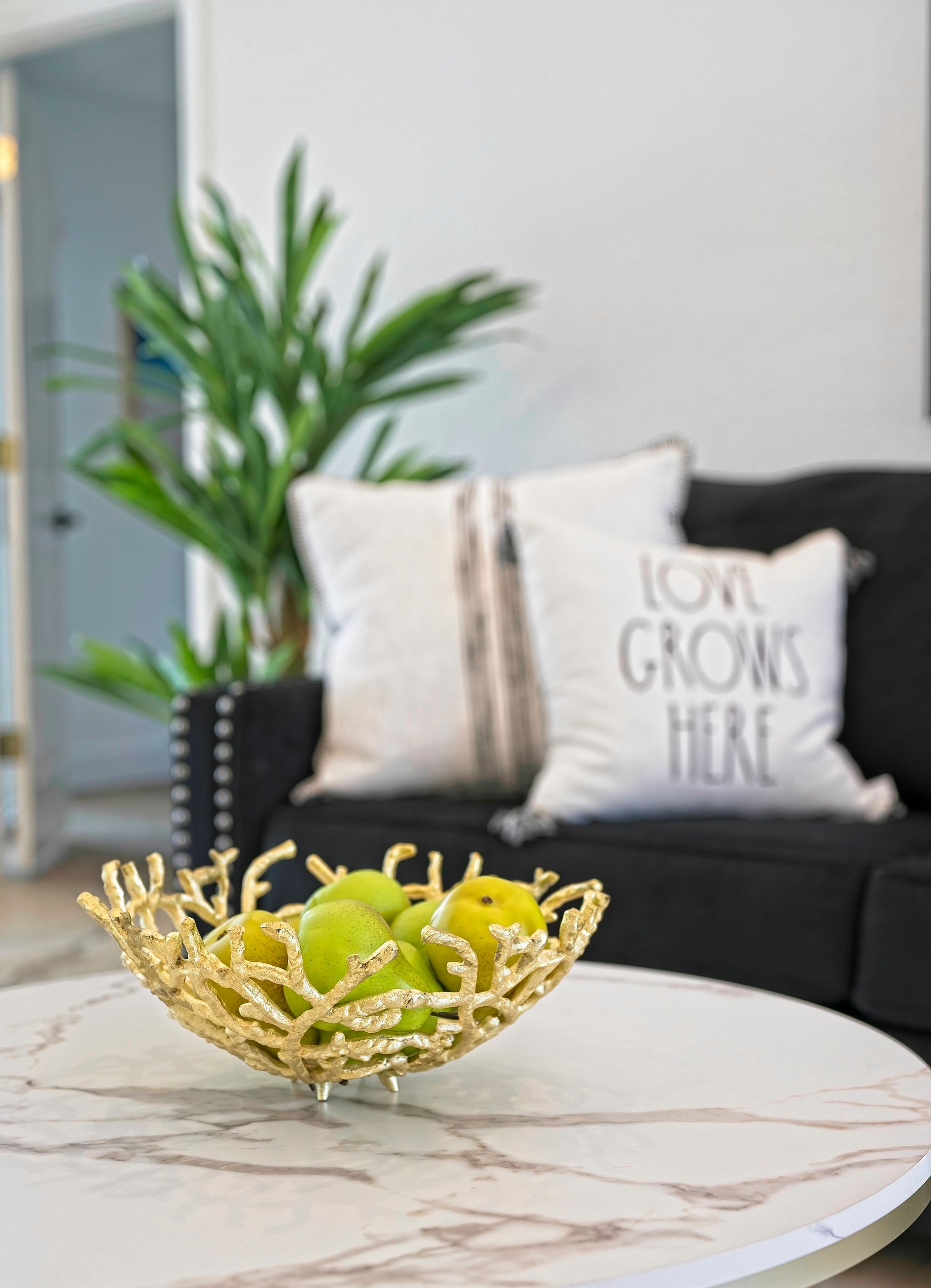 Golden fruit bowl with green apples on a marble coffee table in front of a black couch and pillows.