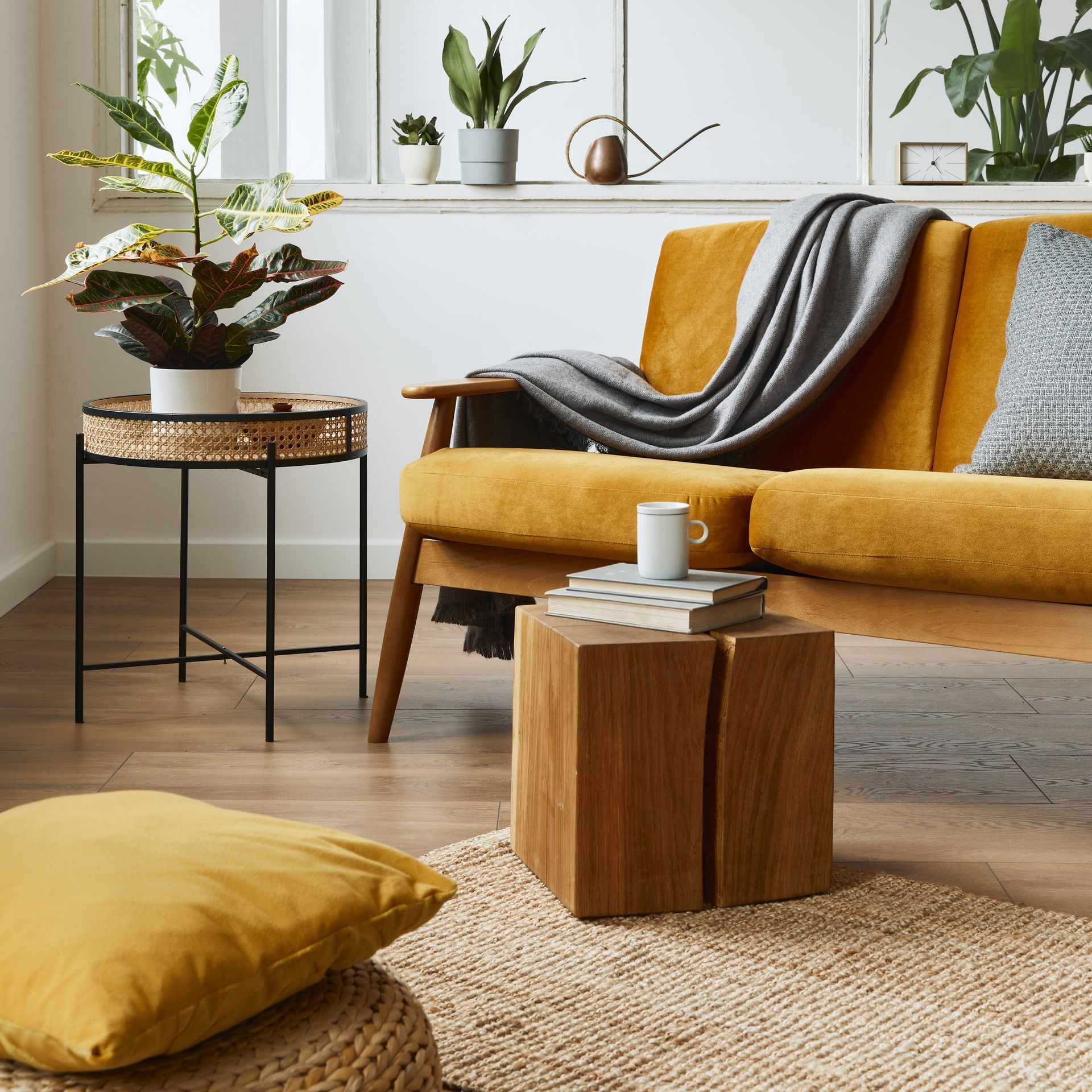 Yellow sofa with throw pillows and blanket, side table with plant, wooden stool, and floor cushion in a bright living room.