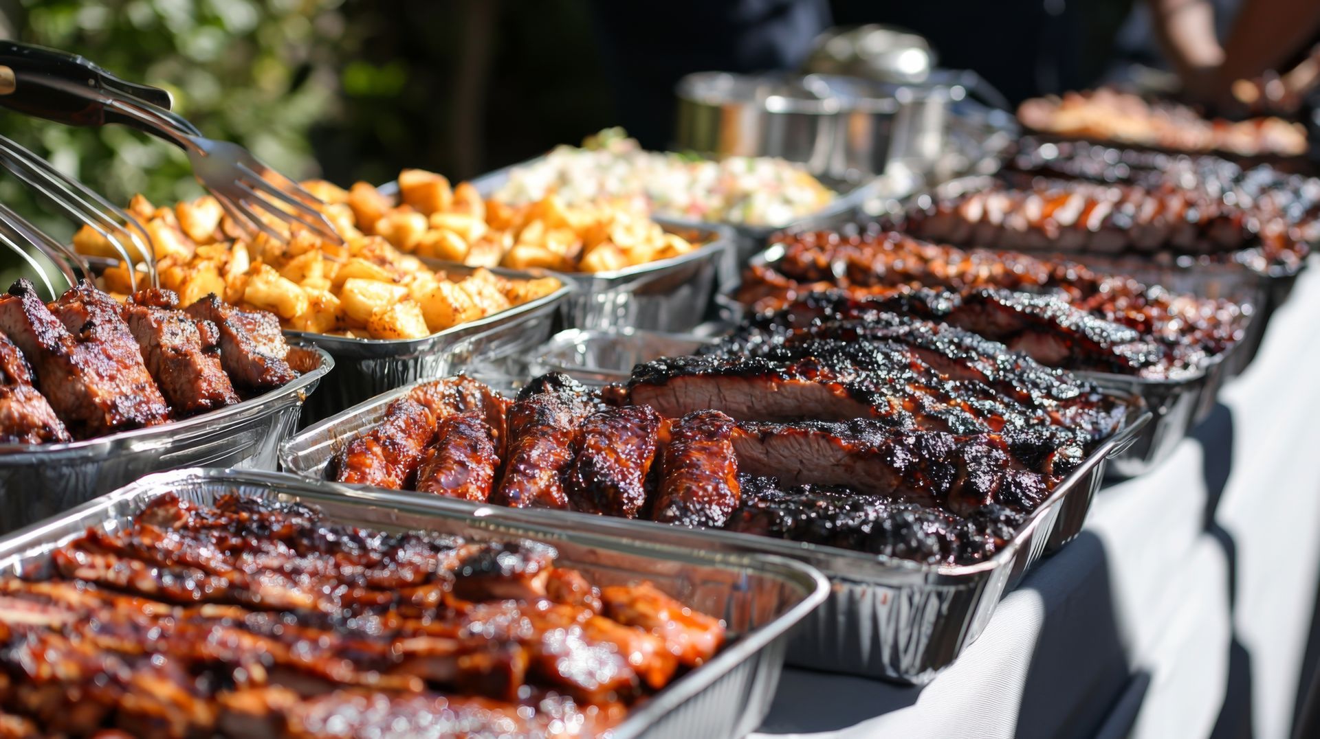 Foil trays filled with grilled barbecue ribs, brisket, and roasted potatoes are arranged on a white buffet table.