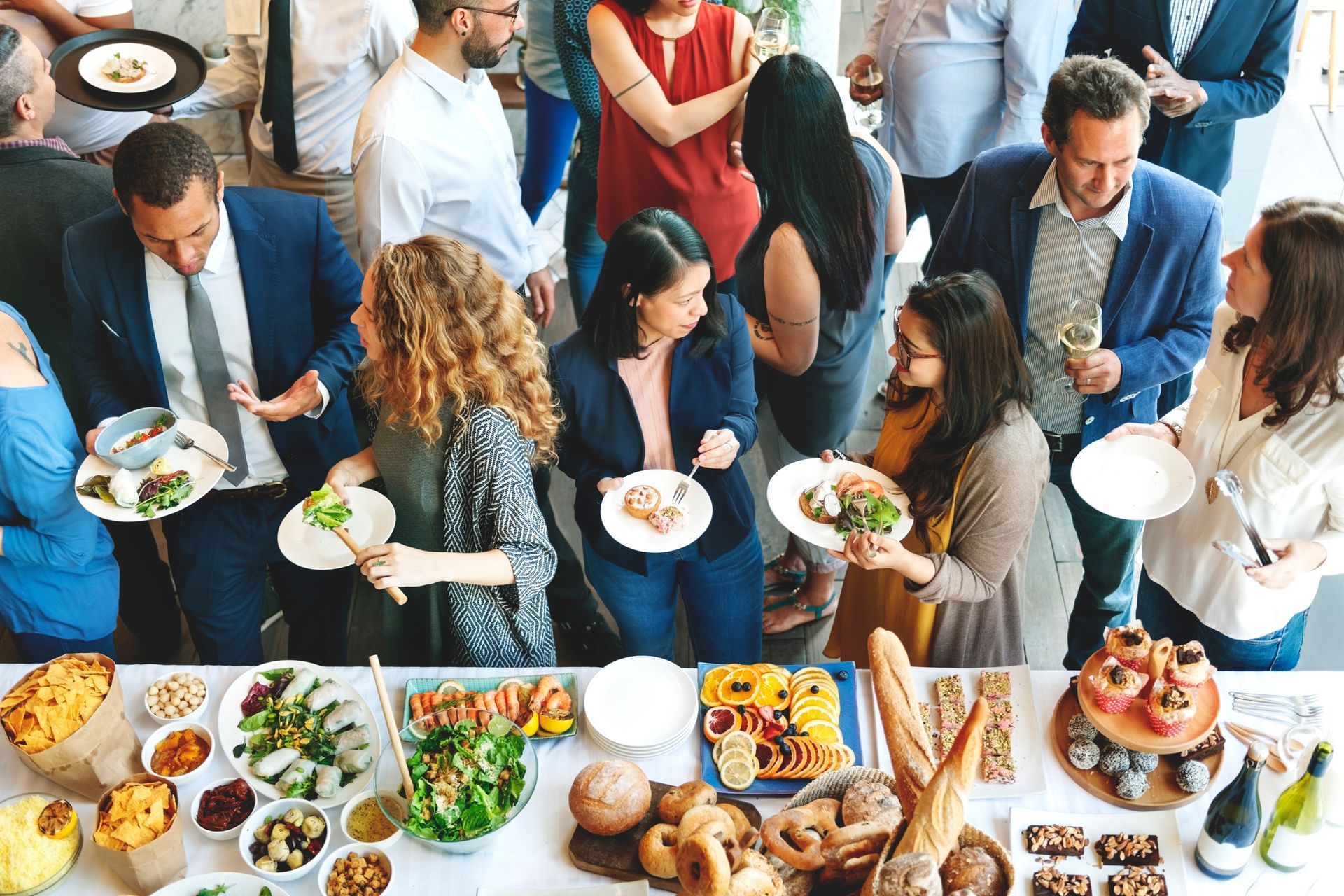 A group of people at an indoor gathering serving themselves from a table laden with food and drinks.