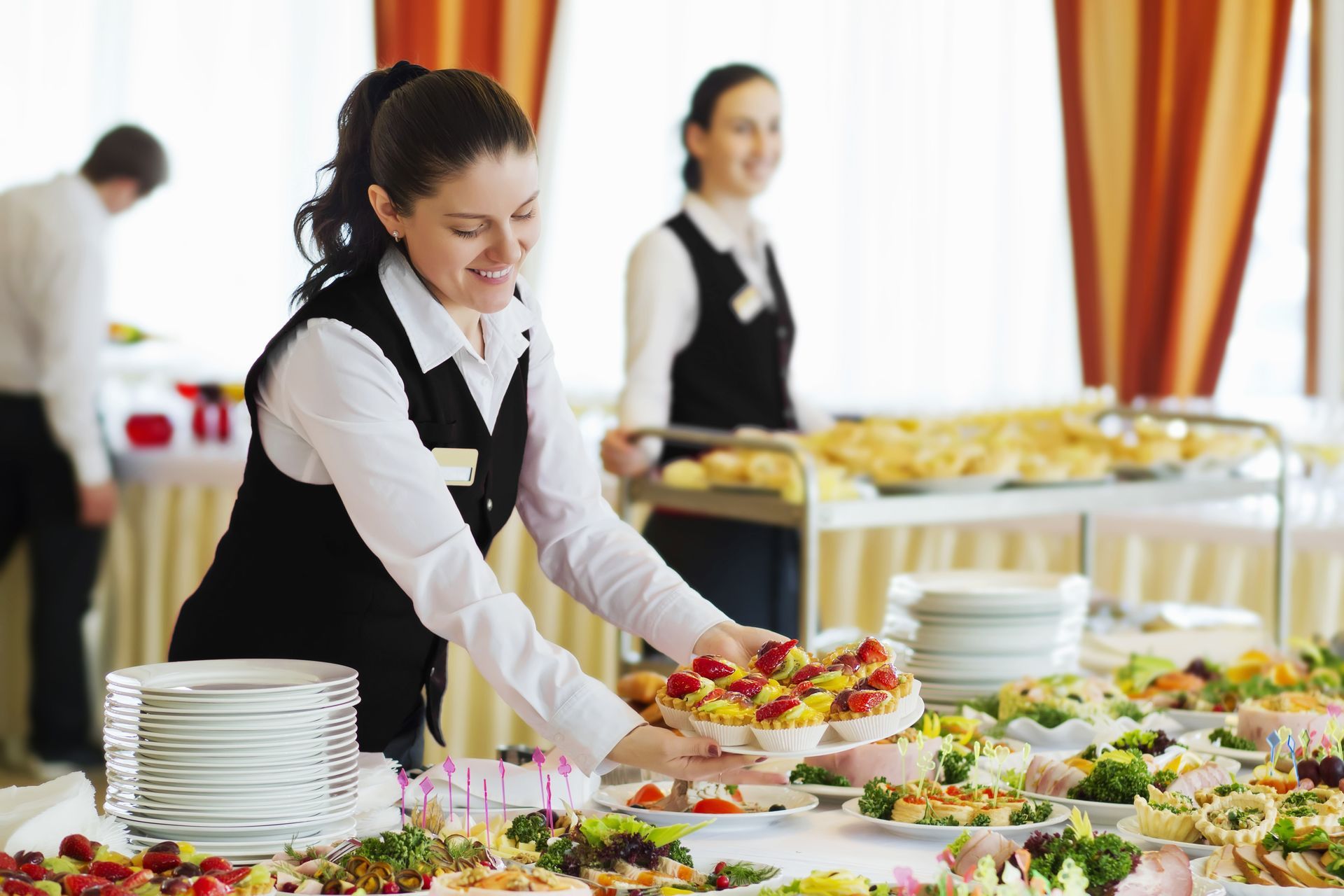 Catering staff in uniform arranging a buffet table with various finger foods and a stack of white plates in a dining hall.