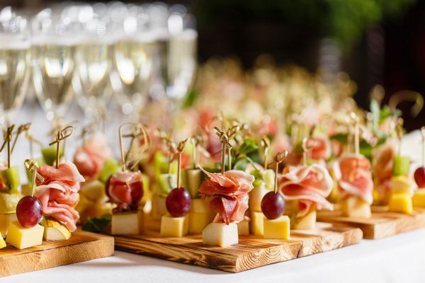 Appetizers on skewers, featuring prosciutto, grapes, and cheese cubes, arranged on wooden boards with glasses in the back.