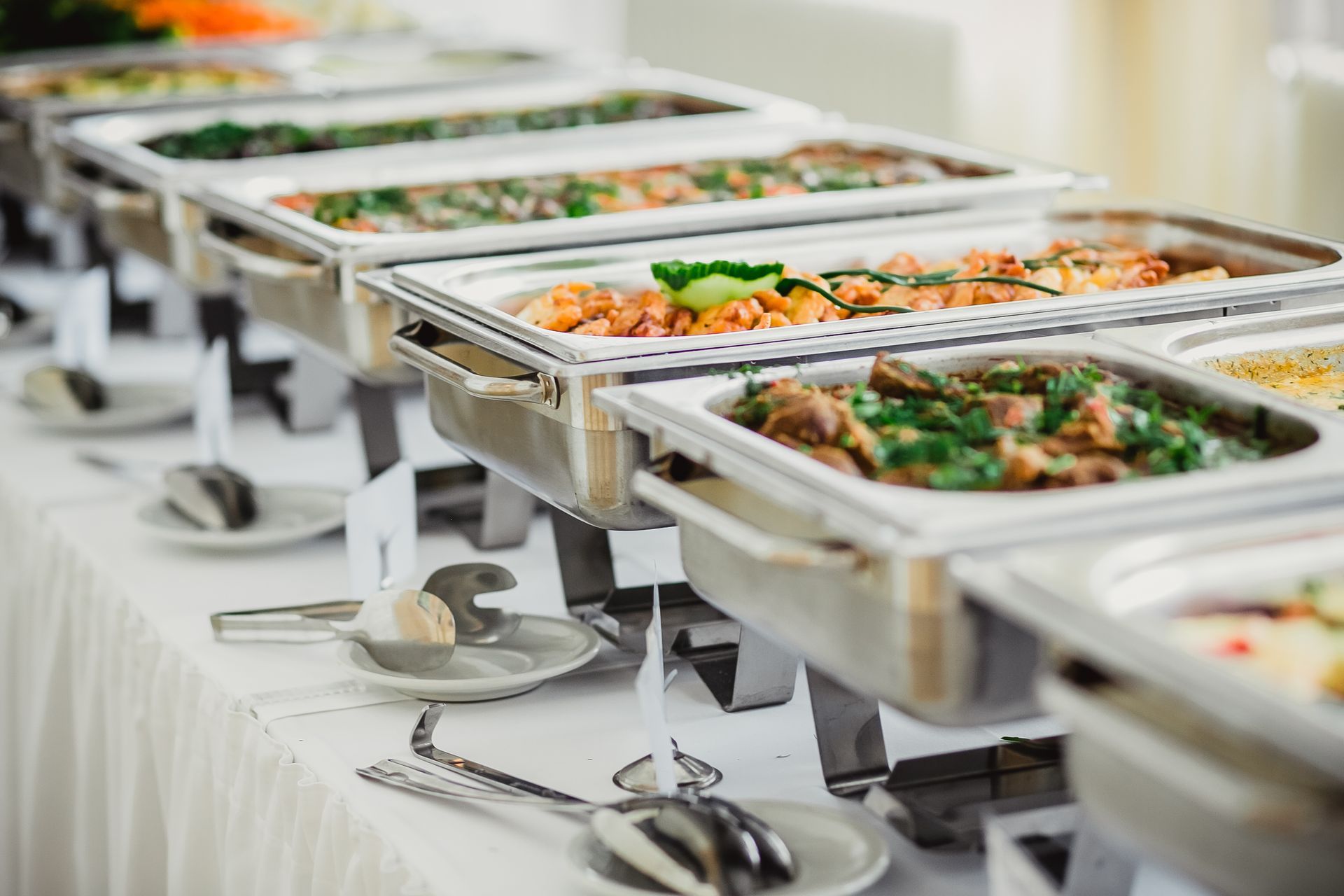 A line of silver buffet chafing dishes filled with various prepared foods arranged on a white tablecloth.