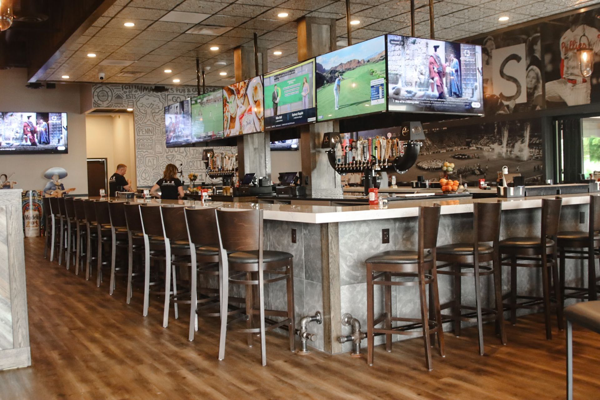 Bar with many TVs showing sports, long countertop with barstools, two bartenders, and decorative wall.