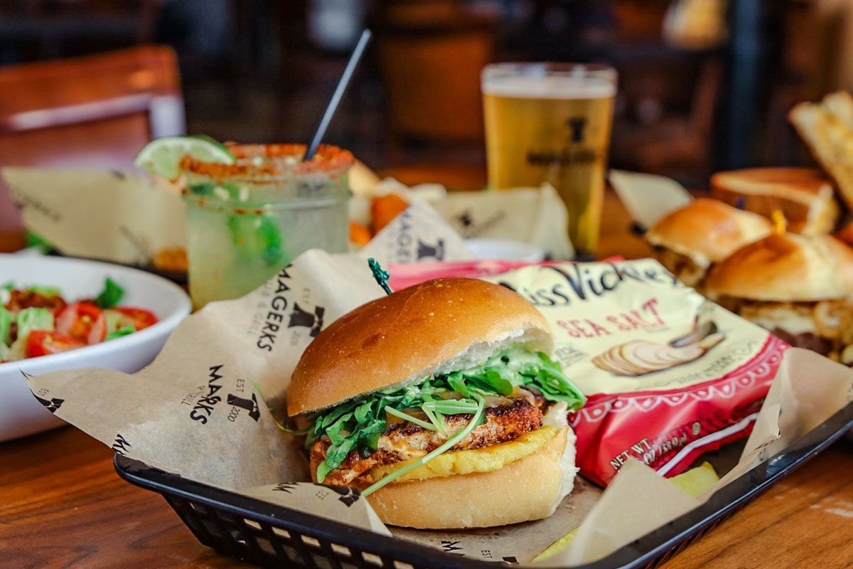 Burger on a tray with chips, drink, and other food on a wooden table.