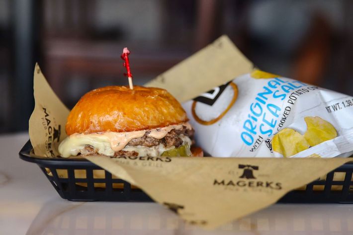 Burger with toothpick, pickles, and sauce in a black basket next to a bag of chips with blue and white packaging.