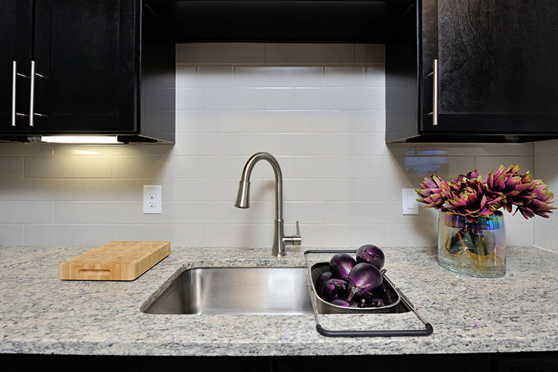 Modern apartment kitchen sink with curved faucet, granite counter, cutting board, purple onions, and a vase of flowers.